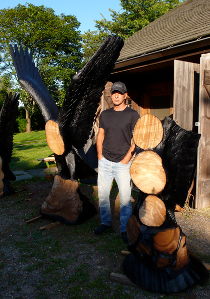 Designer Helmut Lang standing next to a large wooden sculpture of an eagle with outstretched wings outdoors.
